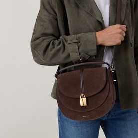 Model carrying DeMellier Vancouver Saddle Suede Crossbody Bag in tan, gold-tone lock charm, adjustable strap, styled with olive jacket and jeans.