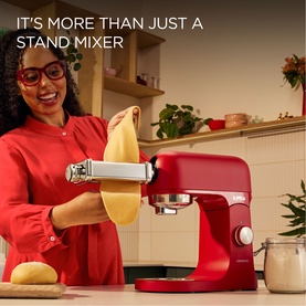 Red stand mixer with pasta roller attachment in use, woman feeding fresh pasta dough through on kitchen counter