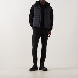 Model wearing Calvin Klein Tech Stretch Slim-Leg Trousers in black, styled with a dark quilted gilet, black hoodie and chunky boots.