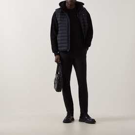 Model wearing Calvin Klein Tech Stretch Slim-Leg Trousers in black, styled with a dark padded gilet, black hoodie, and black leather shoes.