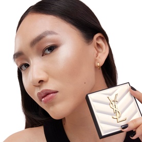Model wearing YSL All Hours Hyper Luminize highlighter, holding gold YSL logo charm in gift box, glowing skin finish visible.