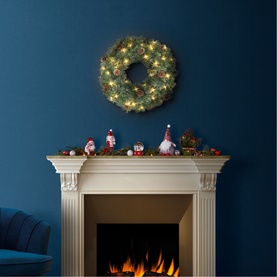 Puleo Pre-lit LED Cashmere Pinecone Christmas Wreath with warm white lights, hung above a fireplace decorated with festive figurines