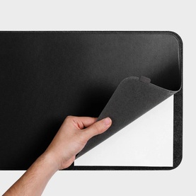 Orbitkey Desk Mat Slim in black faux leather, hand lifting corner to reveal anti-slip white backing