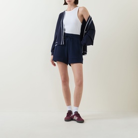Model wearing Varley navy shorts with white vest and matching navy zip-up jacket, styled with burgundy trainers and white socks