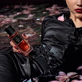 Model with YSL Le Vestiaire des Parfums Jumpsuit Eau de Parfum, holding dark amber glass bottle, wearing black satin outfit with red floral backdrop