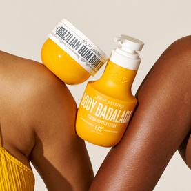Sol De Janeiro Body Badalada Vitamin-Infused Lotion pump bottle and Brazilian Bum Bum Cream jar in yellow, held against tanned skin.