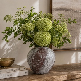 Rustic textured round vase in mottled grey and brown tones, styled with green globe flowers and foliage on a wooden surface.