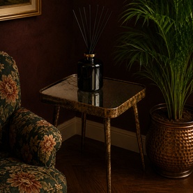 Mindy Browne Ain&eacute; Side Table in gold brown with glass top and slim iron legs, styled with reed diffuser and floral armchair nearby.