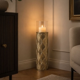 Tall gold textured leaf-pattern pillar candle holder with glass hurricane top, lit candle inside, styled in a warm living room corner.