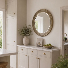 Mindy Browne Elena Round Mirror in gold, oval-shaped with ornate frame, styled above a marble-topped cream cabinet with vase and fruit bowl