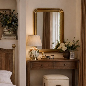 Mindy Browne Isla Curved Mirror in gold, styled above a wooden dressing table with lamp, florals and beauty accessories in a warm bedroom setting.