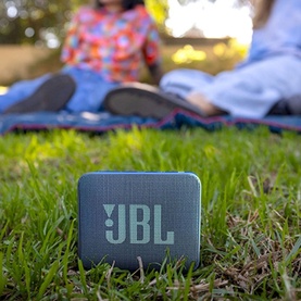 JBL Go 2 Essential portable Bluetooth speaker in blue, placed on grass in outdoor lifestyle setting with people relaxing in background