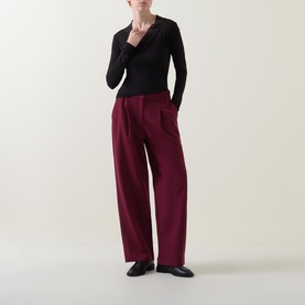 Model wearing Masai Dionius Lace Trim Top in black with V-neckline and long sleeves, styled with wide-leg burgundy trousers and black loafers