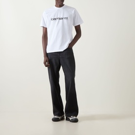 Model wearing Carhartt Script Logo T-Shirt in white with black logo, styled with dark wide-leg jeans and grey trainers.