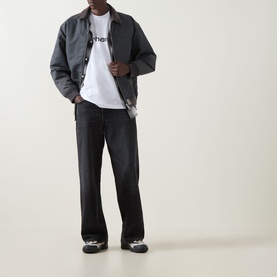 Model wearing Carhartt Script Logo T-Shirt in White/Black with grey coach jacket, black wide-leg jeans and trainers.