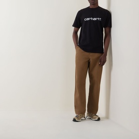 Model wearing Carhartt Script Logo T-Shirt in black with white logo, styled with tan chinos and grey trainers.