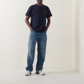 Model wearing Carhartt Chase Logo T-Shirt in navy, crew neck with gold chest logo, styled with mid-wash jeans and grey trainers.