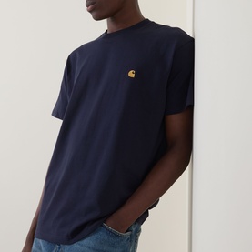 Model wearing Carhartt Chase Logo T-Shirt in navy, crew neck with gold embroidered chest logo, styled with light blue jeans.