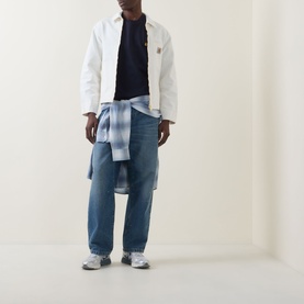 Model wearing Carhartt Chase Logo T-Shirt in Navy, styled with white jacket, blue check shirt tied at waist, and light wash jeans.