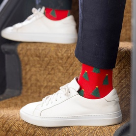 Model wearing Swole Panda Christmas Tree Socks in red with green tree print, styled with white leather trainers and navy trousers