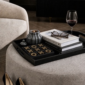 Black decorative tray styled with noughts and crosses game, stacked books, glasses, candle holder and wine glass on cream boucle ottoman