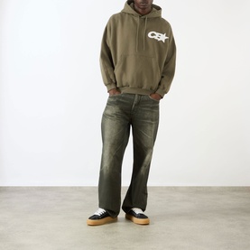 Model wearing Cole Buxton Star Racing Hoodie in khaki with contrast chest logo, styled with distressed wide-leg jeans and low-top trainers.