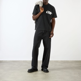Model wearing Cole Buxton Vintage Black cotton T-shirt with logo graphic, styled with black wide-leg trousers and black chunky loafers.