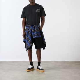 Model wearing Cole Buxton Sportswear Letter Logo T-Shirt in Vintage Black, styled with blue tartan shirt tied at waist and black shorts.