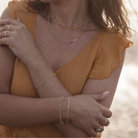 Model with Juvi Bali Beaded Chain Bracelet in gold, styled with layered gold necklaces and rings, wearing a mustard ruffle top