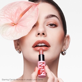 Model wearing NARS Explicit Lipstick in shade Scandal, holding bullet lipstick, styled with pink anthurium hair accessory