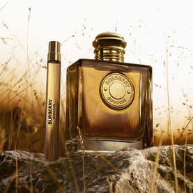 Burberry Goddess Parfum for Women 10ml travel spray alongside full-size amber glass bottle with gold cap, on stone amid golden grass