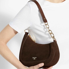 Model carrying Dune Direction Saddle Shoulder Bag in brown suede with gold-tone chain handle, worn on shoulder, styled with white top