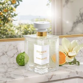 Balmain Blanc Galaxie Eau de Toilette clear glass bottle with gold cap on marble surface, styled with citrus fruits and white flower