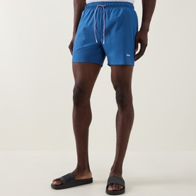 Model wearing Hugo Boss blue swim shorts with contrast drawstring, styled with dark slides.