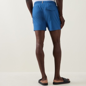 Model wearing Hugo Boss blue swim shorts, rear view, with black slider sandals. Elasticated waistband and back pocket visible.