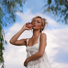 Model with OREBELLA Eternal Roots Parfum 50ml, holding bottle near face, wearing white lace dress, outdoor sky backdrop