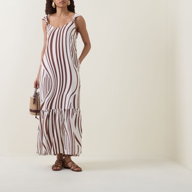 Model wearing Seafolly Striped Tiered Dress in Cocoa, maxi length with tiered skirt and knotted straps, styled with brown sandals and small bag.