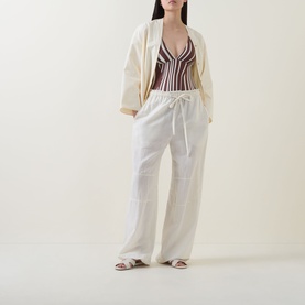 Model wearing Seafolly swimsuit in Cocoa with vertical stripe detail, styled with white wide-leg trousers, cream blazer and tan sandals.