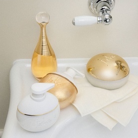 Dior J'adore Les Adorables Shimmering Oil displayed on a bathroom sink edge with matching gold and white body care accessories