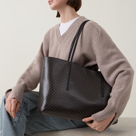 Model carrying Calvin Klein Monogram Logo Tote Bag in black with all-over logo pattern and carry handles, styled with beige knit jumper and jeans