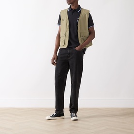 Model wearing Barbour Lure Logo Polo Shirt in Dark Navy, styled with a beige gilet, black trousers and black high-top trainers.