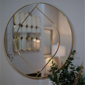 Round wall mirror with gold geometric metal frame, hung above decorative greenery in a styled living space