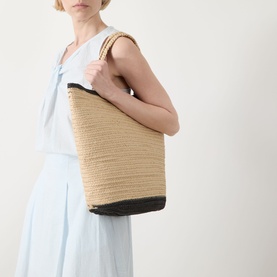 Model carrying Part Two Biggie Woven Tote Bag in natural paper weave with contrast dark trim and short top handles, styled with light blue dress