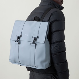Model carrying Rains MSN Waterproof Backpack in pool blue, with carabiner closure and adjustable straps, worn over black puffer jacket.