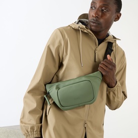 Model carrying Rains Bum Bag in olive green, worn crossbody over a tan hooded jacket with black strap visible.