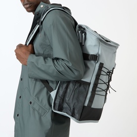 Model carrying Rains Trail Mountaineer Bag in Pool, a structured roll-top backpack with front bungee cord storage and side mesh pocket.