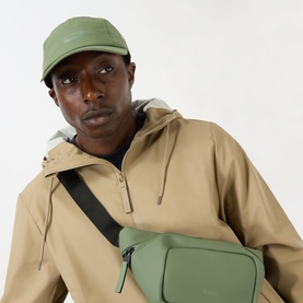 Model with Rains 5 Panel Water-Repellent Cap in green, styled with matching green crossbody bag and tan hooded jacket.