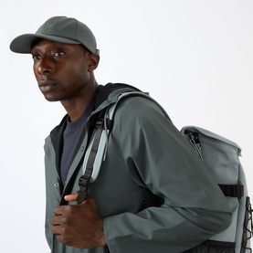 Model with Rains Water-Repellent Cap in dark grey, six-panel curved brim style, styled with matching grey jacket and backpack.