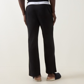 Model wearing Calvin Klein black cotton lounge bottoms, rear view, with logo waistband and wide-leg fit, styled with tan slippers.