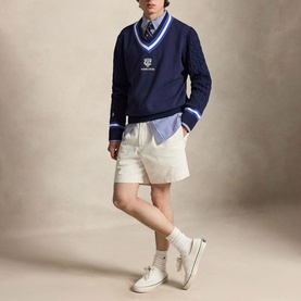 Model wearing Ralph Lauren Wimbledon Logo Hybrid Sweater in Refined Navy with striped V-neck, styled with cream shorts and white trainers.